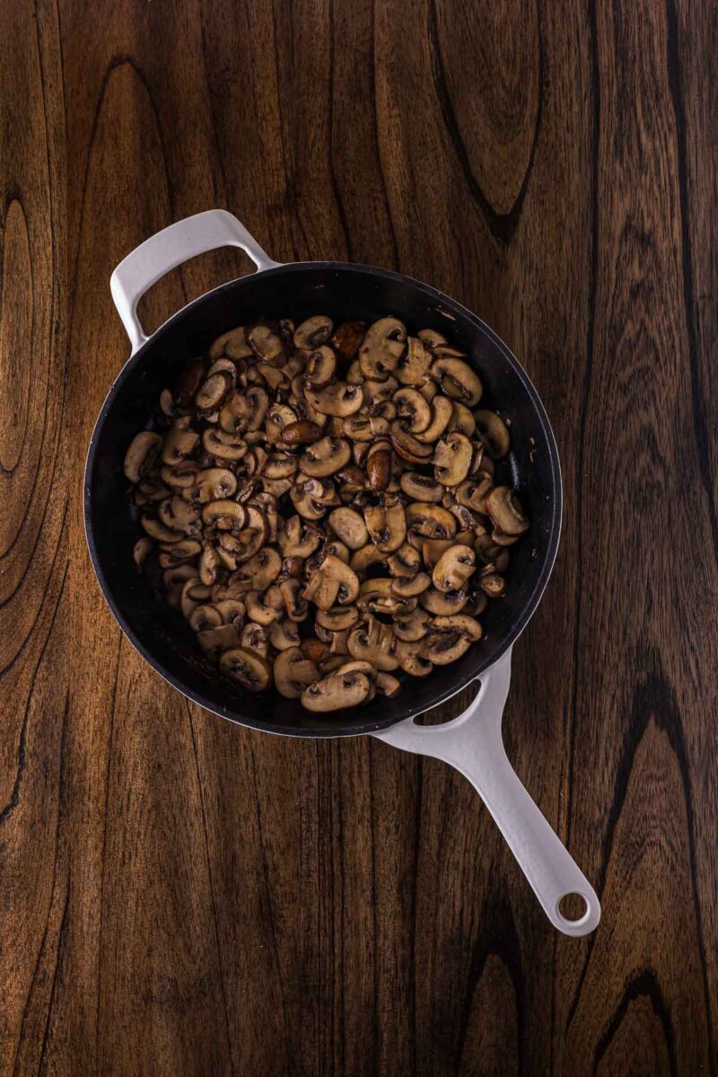 A white-handled skillet filled with sautรฉed sliced mushrooms sits on a dark wooden surface.