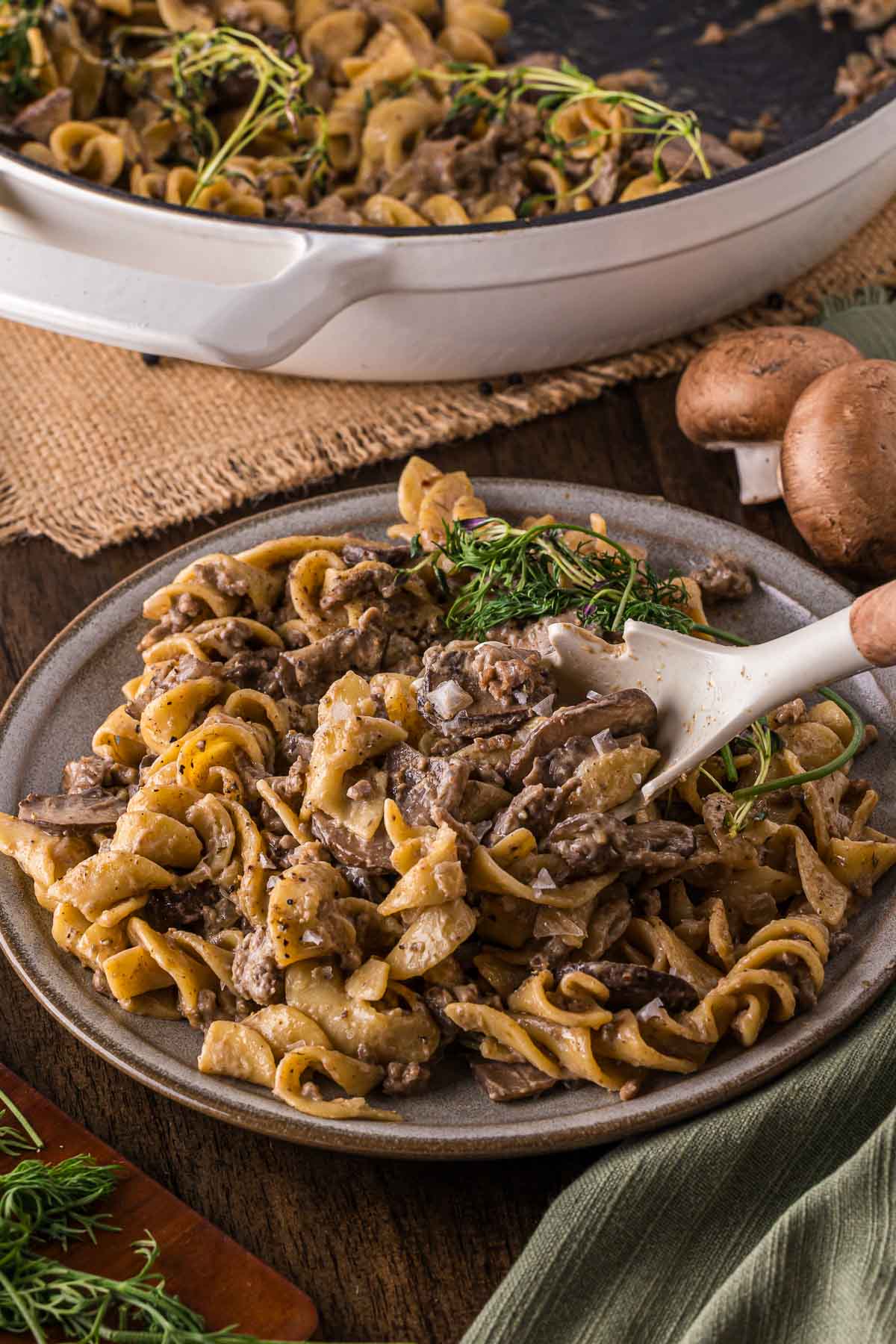 A plate of beef stroganoff with ground beef with egg noodles is served with a sprig of herbs, next to a white skillet and fresh mushrooms on a rustic wooden table.