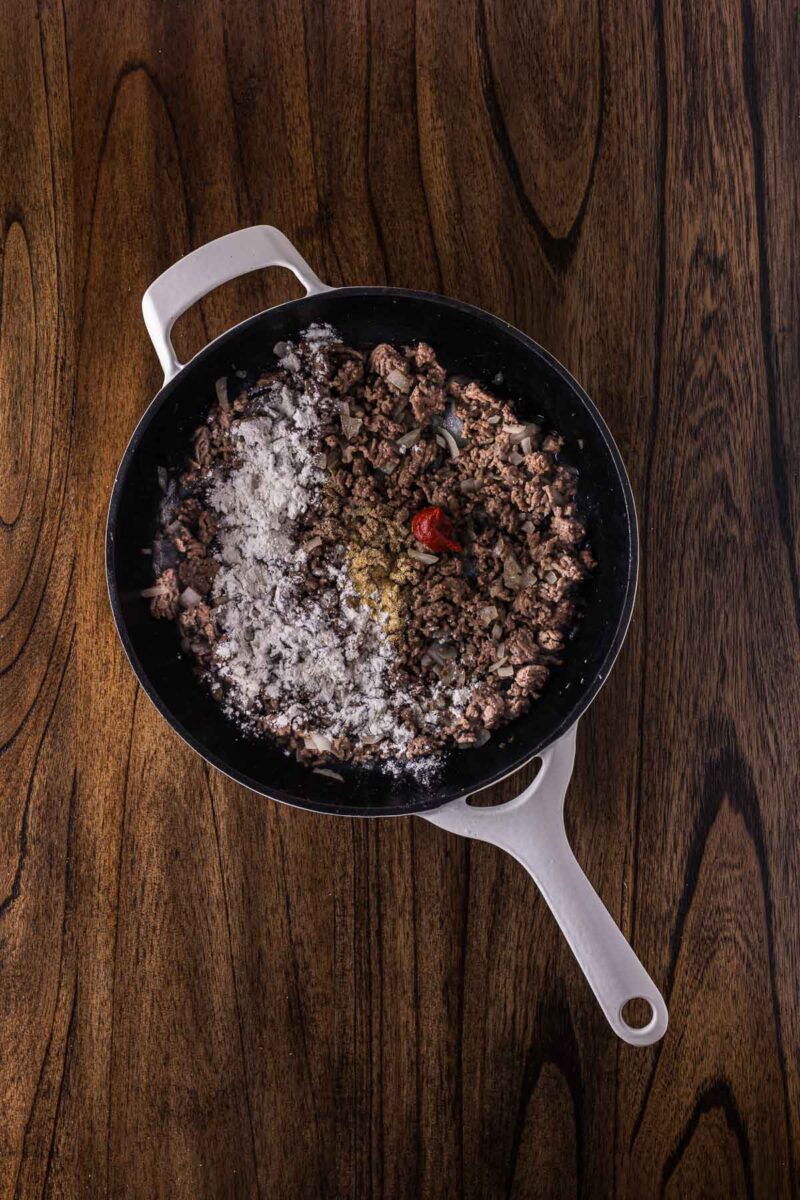 A white-handled skillet containing ground meat with seasonings and flour on top, placed on a wooden surface.