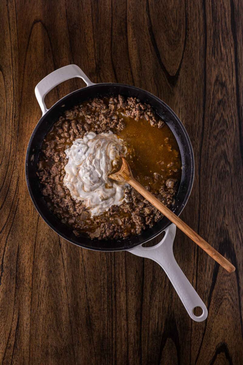 A white cast iron skillet with cooked ground beef, brown sauce, and a dollop of creamy mixture being stirred with a wooden spoon on a wooden surface.