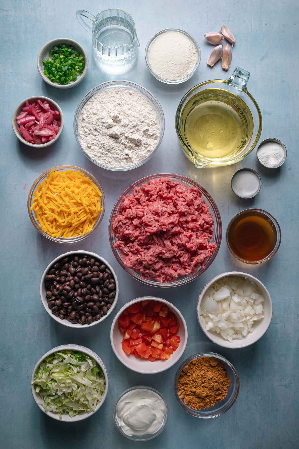 Various taco ingredients arranged on a blue surface, including ground beef, shredded cheese, flour, beans, onions, peppers, lettuce, seasonings, oil, sour cream, broth, and water.