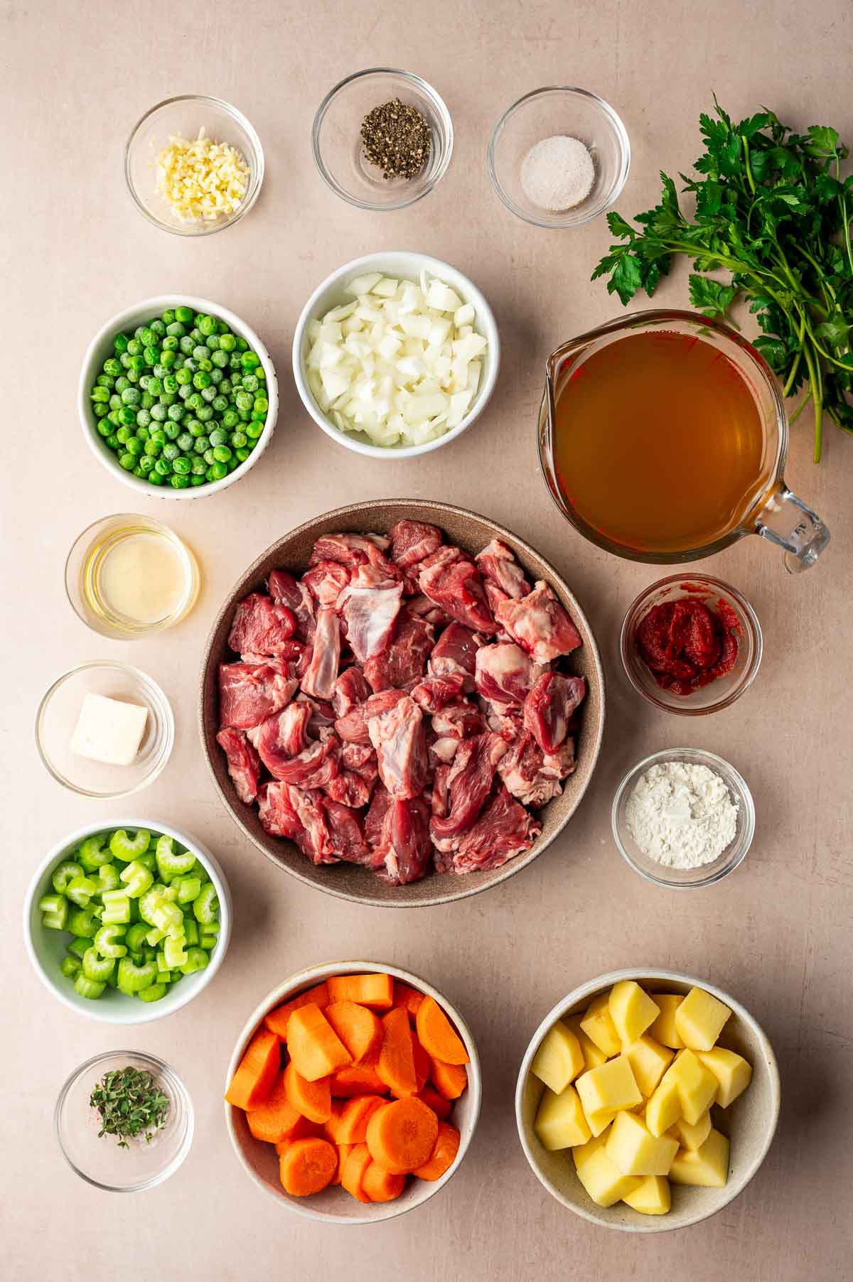 Raw stew ingredients arranged on a table, including cubed lamb shoulder, carrots, potatoes, celery, peas, onions, broth, tomato paste, garlic, herbs, and seasonings in separate bowls.