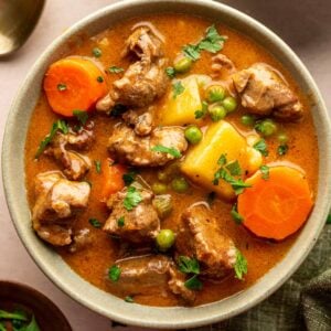 A bowl of lamb stew with chunks of lamb, carrots, potatoes, peas, and fresh parsley garnish, served on a light surface with a spoon nearby.