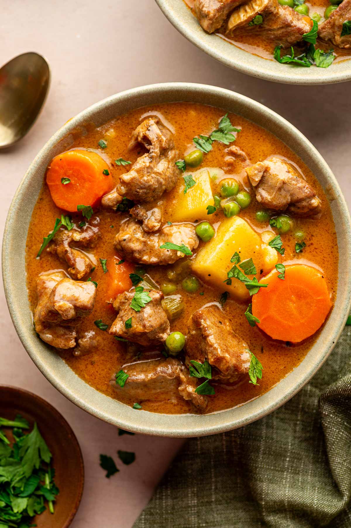 A bowl of lamb stew with chunks of lamb, carrots, potatoes, peas, and fresh parsley garnish, served on a light surface with a spoon nearby.