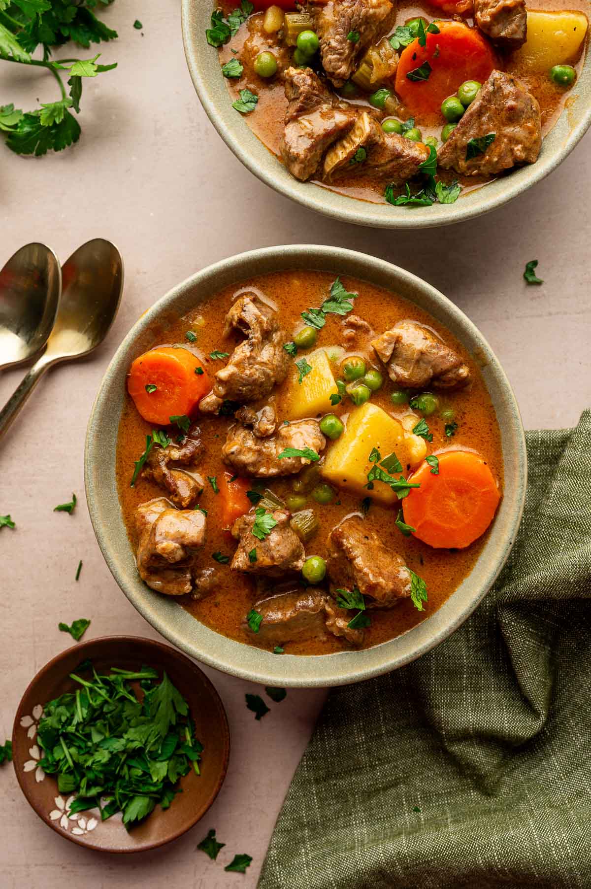 Two bowls of lamb stew with carrots, potatoes, peas, and fresh herbs, placed on a table with a green cloth, a small plate of chopped parsley, and two spoons.