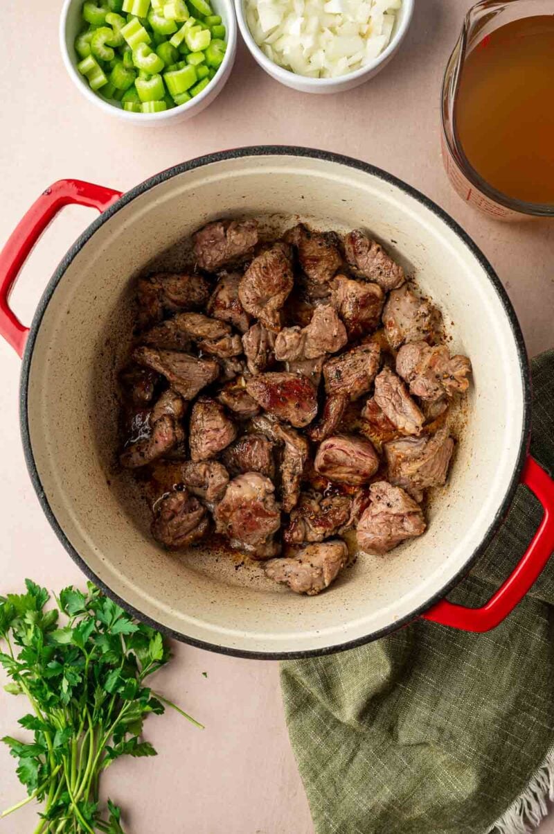 Chunks of browned lamb in a red Dutch oven, surrounded by chopped celery, onions, parsley, and a measuring cup of broth.