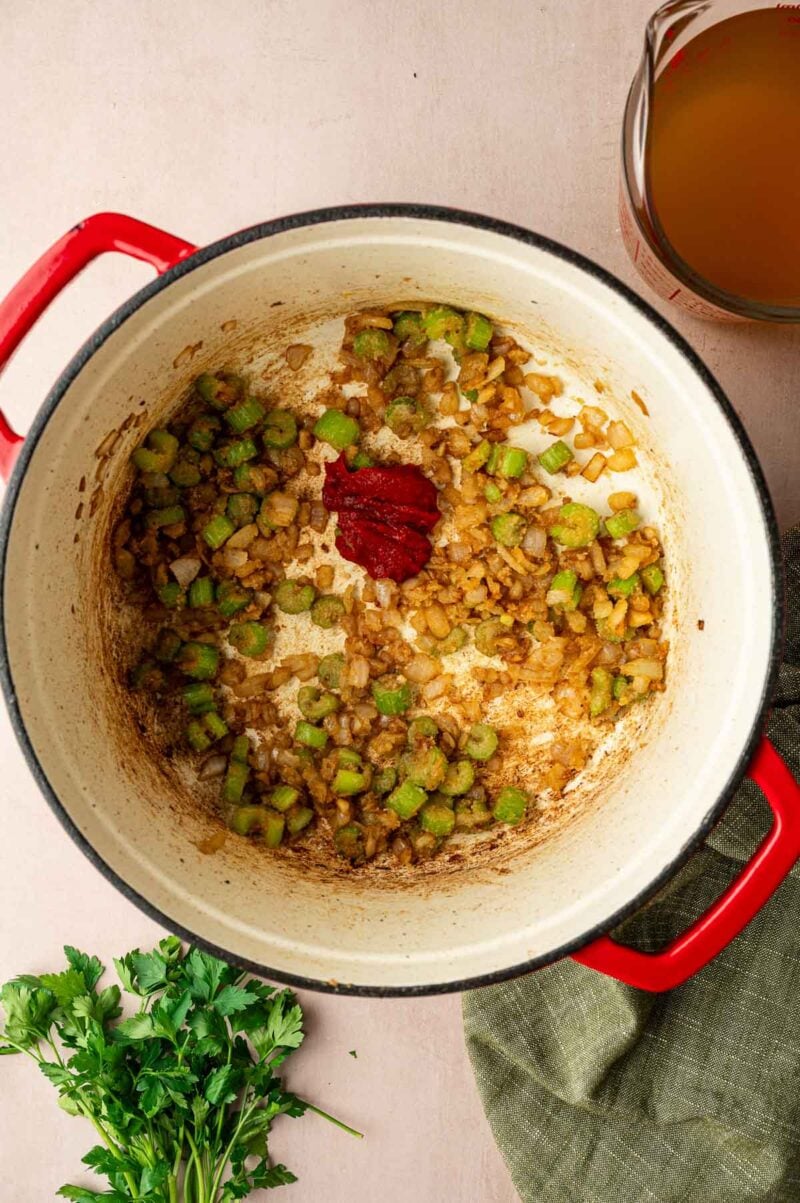 A red Dutch oven with sautéed onions and celery, tomato paste in the center, a cup of broth nearby, fresh parsley, and a green cloth on the side.