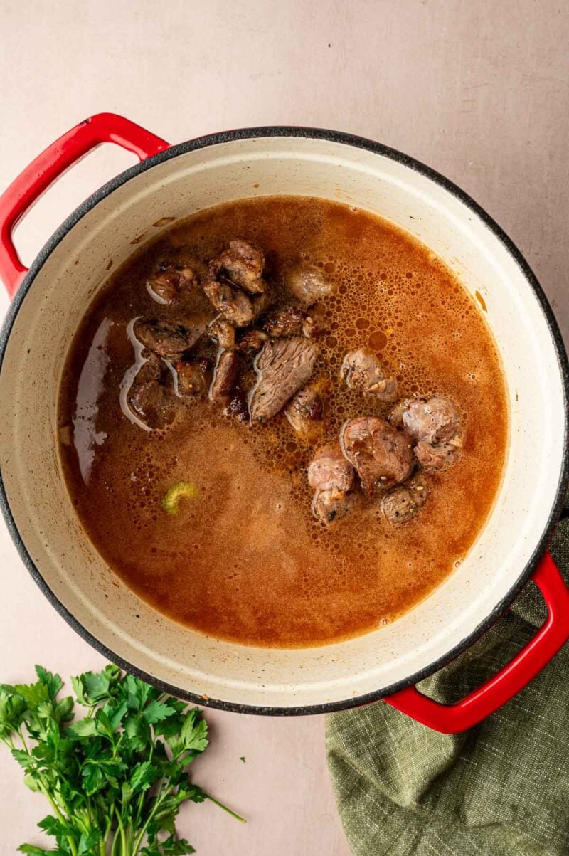 A red-handled pot filled with simmering brown stew and lamb meat, placed on a light surface next to fresh parsley and a green cloth.