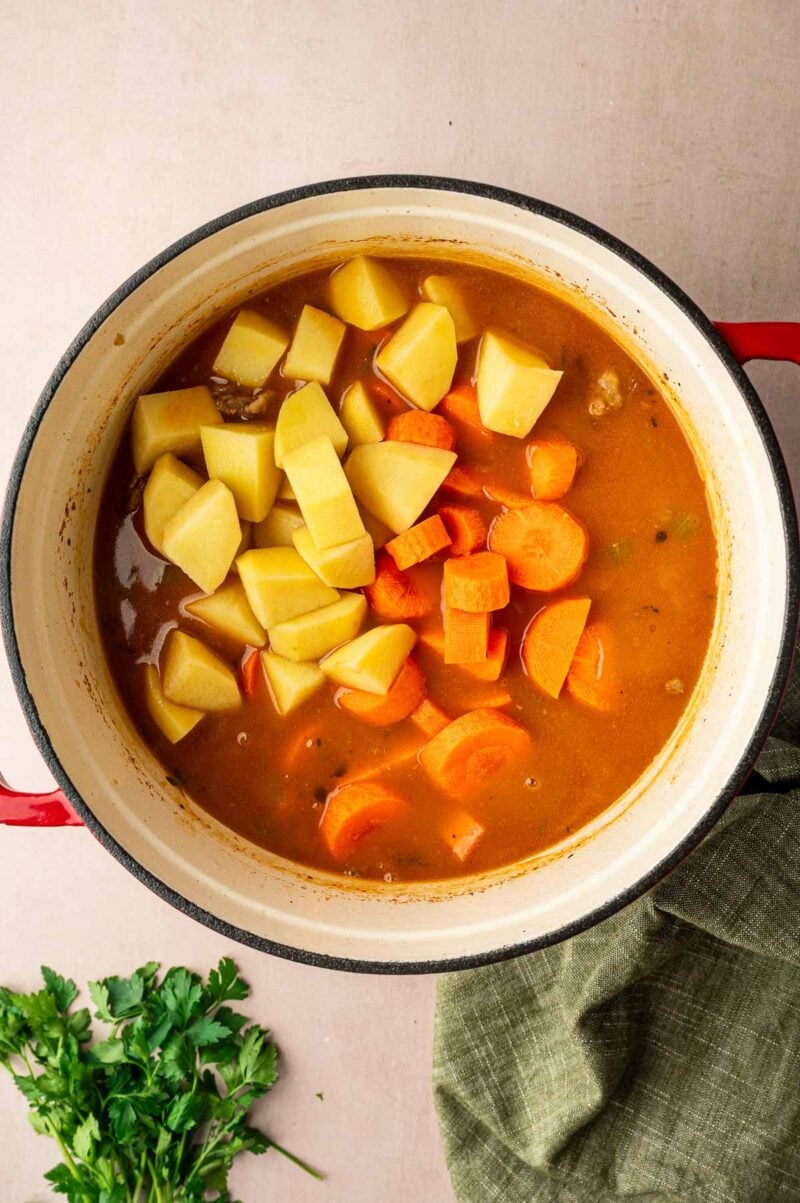 A pot filled with stew containing chunks of potatoes and carrots in a brown broth, placed on a countertop next to a bunch of fresh parsley.