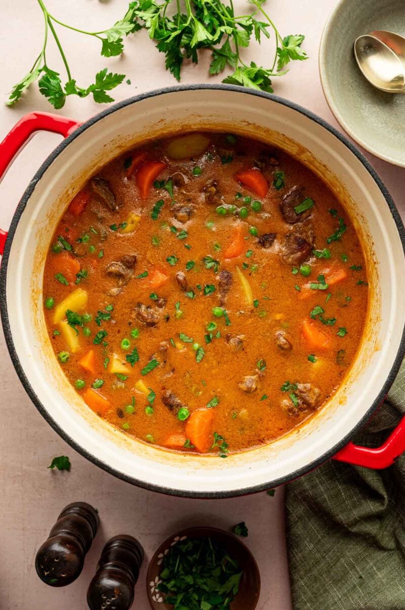 A Dutch oven of lamb stew with carrots, potatoes, and herbs sits on a table next to fresh parsley, a spoon, and pepper grinders.