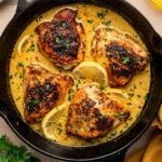 A cast iron skillet with four seasoned chicken thighs in creamy sauce, garnished with lemon slices and herbs, on a wooden surface with parsley and a yellow napkin nearby.