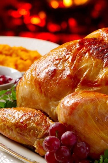 A roasted turkey on a platter with grapes, served alongside mashed sweet potatoes and cranberry sauce, with a warm fire in the background.