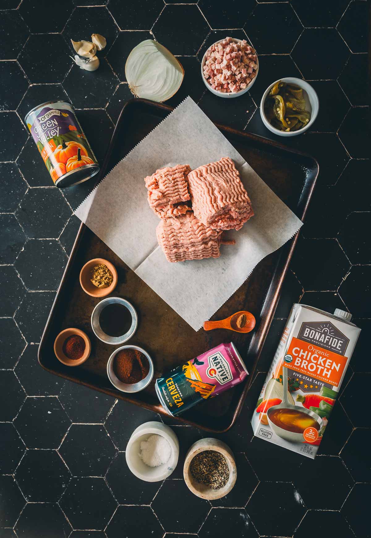 Ground turkey on a tray surrounded by cans, spices, chopped onions, broth, garlic, jalapeños, and seasonings on a black hexagon tile surface.