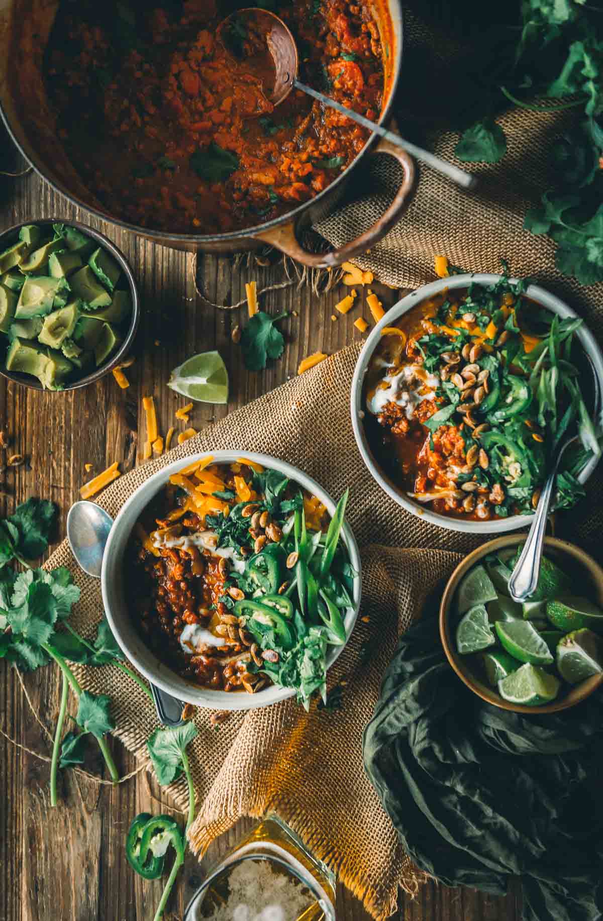 Overhead view of two bowls of pumpkin chili with ground turkley topped with greens, cheese, and sour cream, surrounded by lime wedges, avocado, cilantro, and a pot of chili on a wooden table.