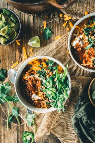 Two bowls of pumpkin chili with ground turkley topped with cheese, sliced green onions, cilantro, and sour cream, with a side bowl of diced avocado and scattered garnishes on a rustic wooden table.