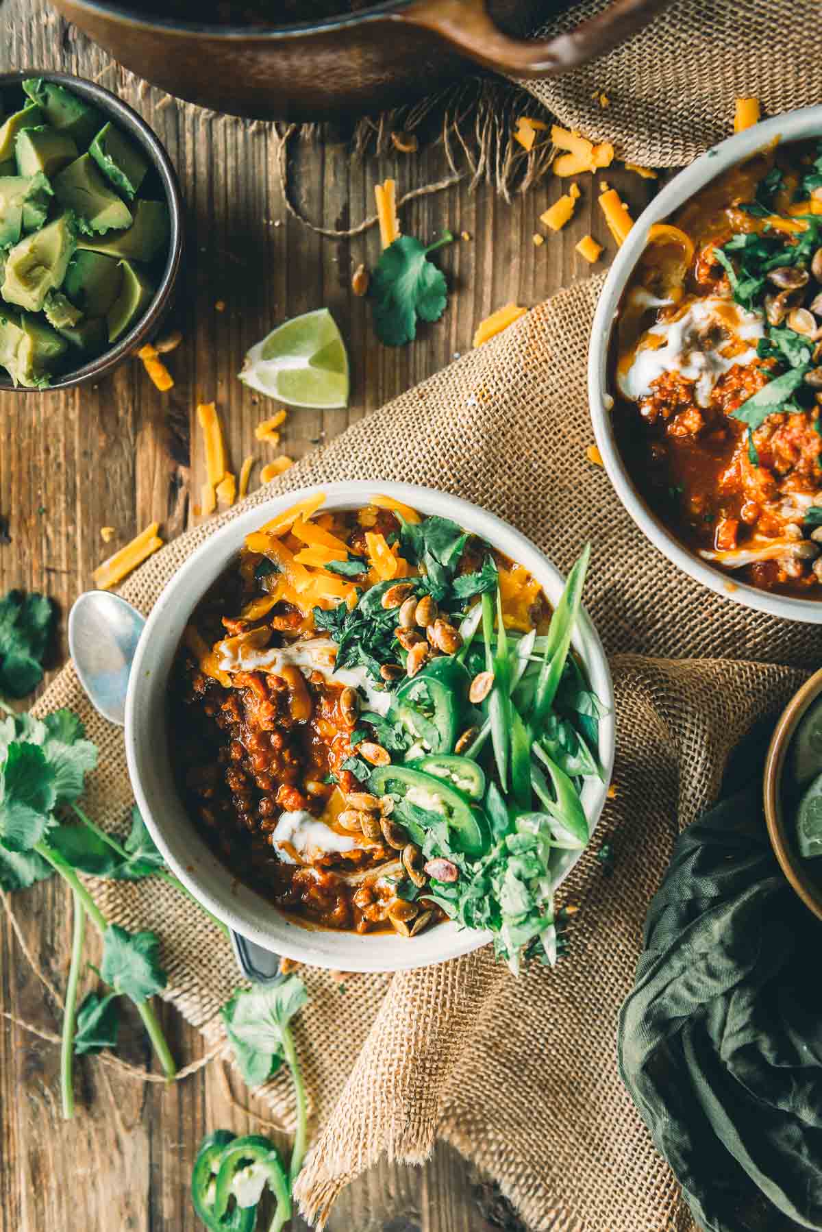 Two bowls of pumpkin chili with ground turkley topped with cheese, sliced green onions, cilantro, and sour cream, with a side bowl of diced avocado and scattered garnishes on a rustic wooden table.