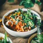 A bowl of pumpkin chili with ground turkley topped with greens, seeds, and herbs, placed on a burlap surface with lime wedges and more greens in the background.