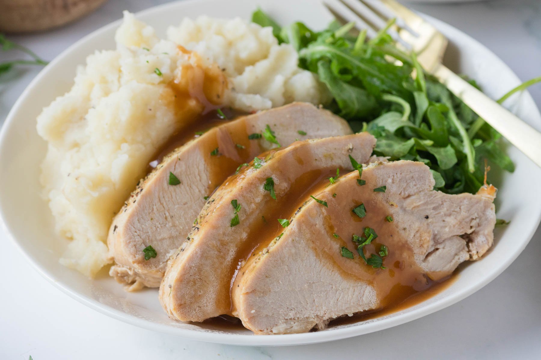 Sliced turkey breast with gravy, mashed potatoes, and a side of greens served on a white plate with a fork.