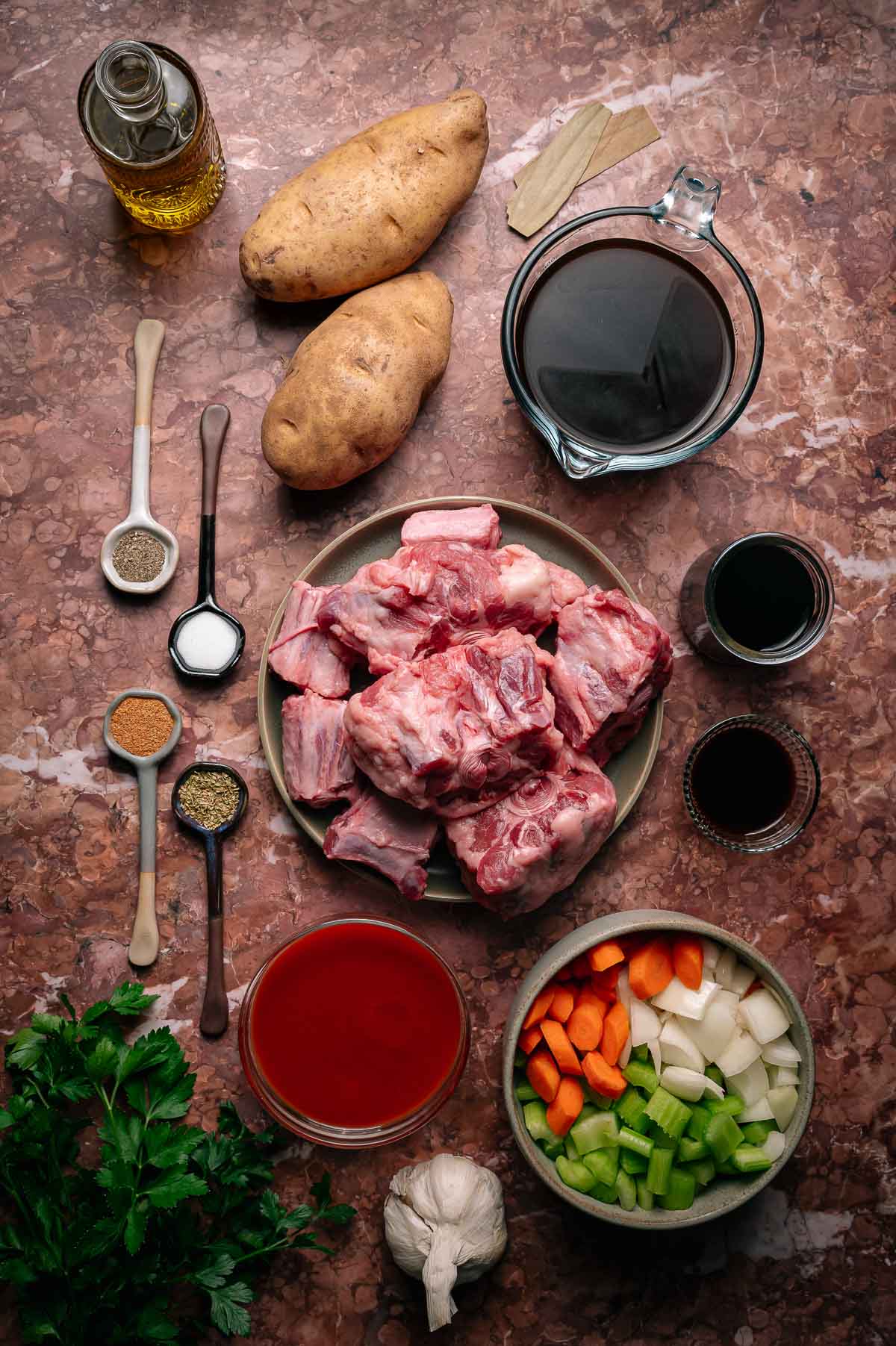 Assorted raw ingredients for oxtail stew, including oxtail pieces, potatoes, chopped vegetables, seasonings, tomato sauce, wine, garlic, oil, and bay leaves, arranged on a marble surface.
