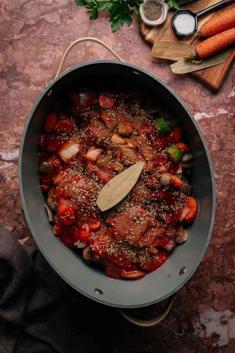 An oval pot filled with chopped vegetables, oxtails, sauce, and a bay leaf sits on a brown countertop next to a cutting board with carrots, seasonings, and herbs.
