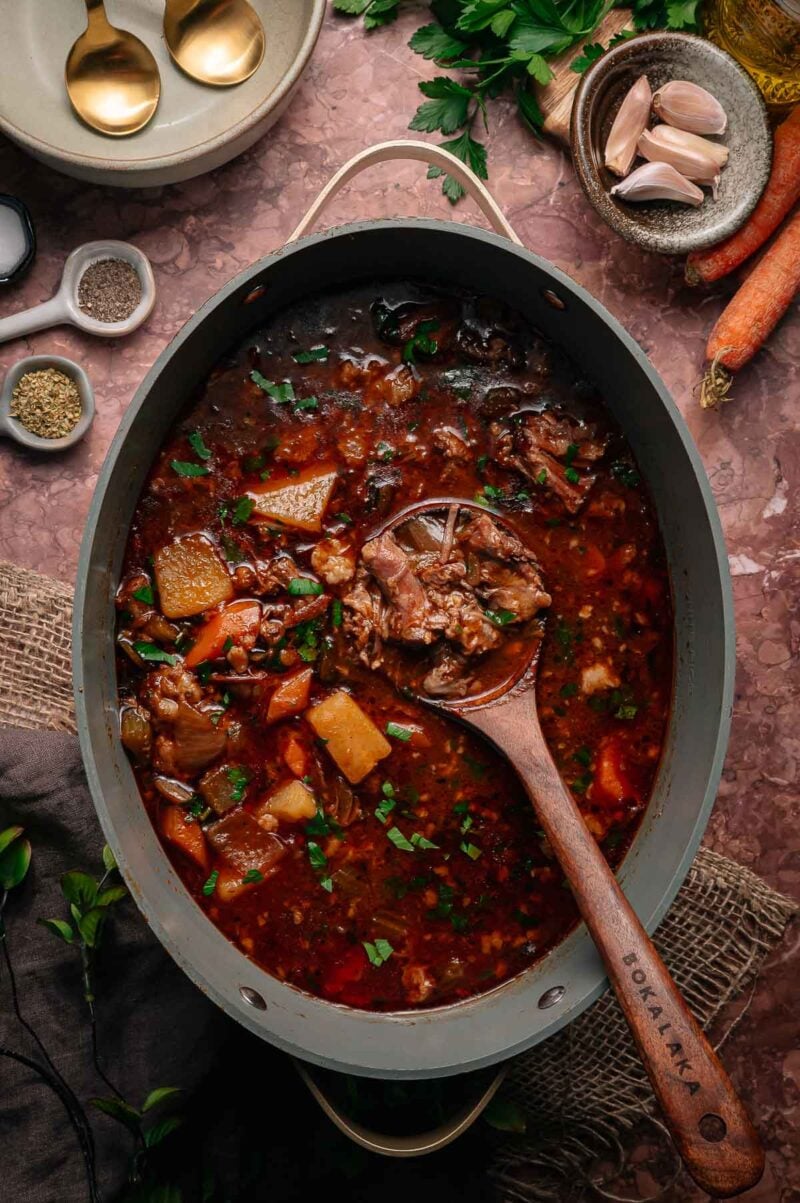 A pot of oxtail soup with chunks of meat, potatoes, and carrots in a rich broth, garnished with chopped parsley. A wooden spoon rests in the stew.