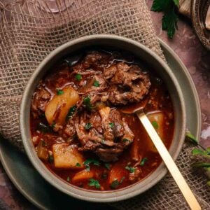 A bowl of oxtail stew with potatoes in a rich, dark broth, garnished with herbs, placed on a rustic mat with a gold spoon. A glass of red wine and fresh herbs are nearby.