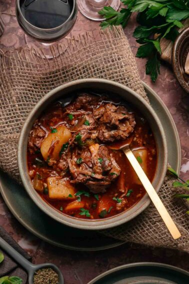 A bowl of oxtail stew with potatoes in a rich, dark broth, garnished with herbs, placed on a rustic mat with a gold spoon. A glass of red wine and fresh herbs are nearby.