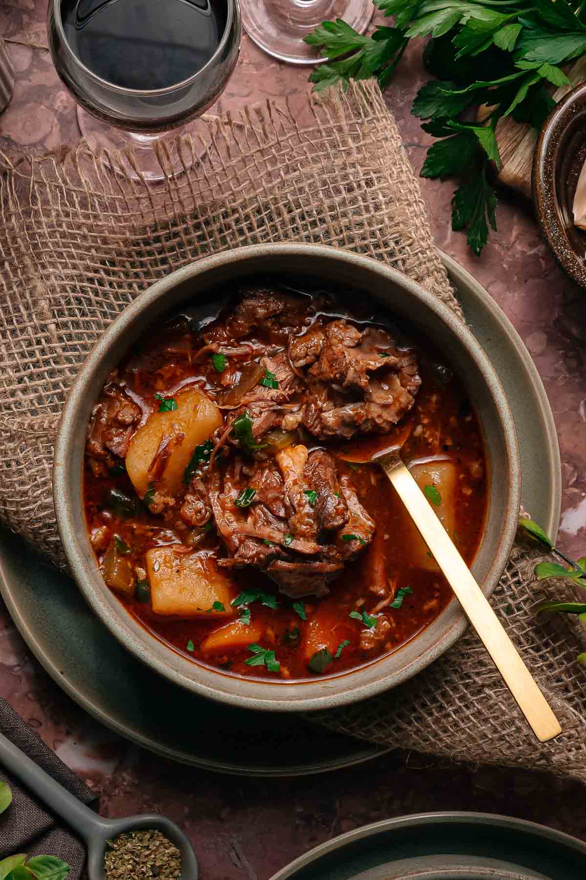 A bowl of oxtail stew with potatoes in a rich, dark broth, garnished with herbs, placed on a rustic mat with a gold spoon. A glass of red wine and fresh herbs are nearby.