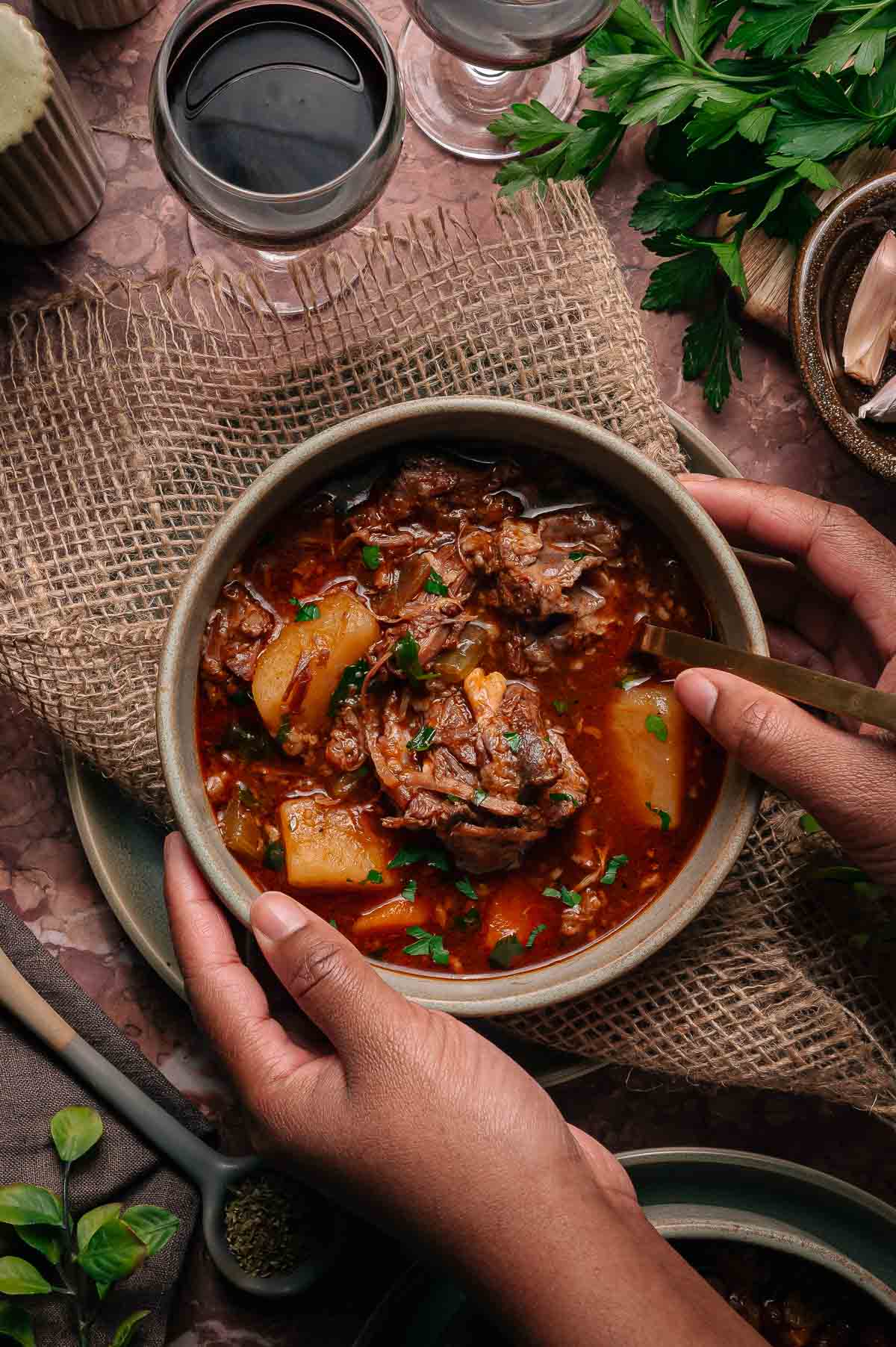 A person holds a bowl of oxtail soup with chunks of shredded beef and potatoes, garnished with herbs, next to a glass of red wine and fresh parsley on a rustic tabletop.