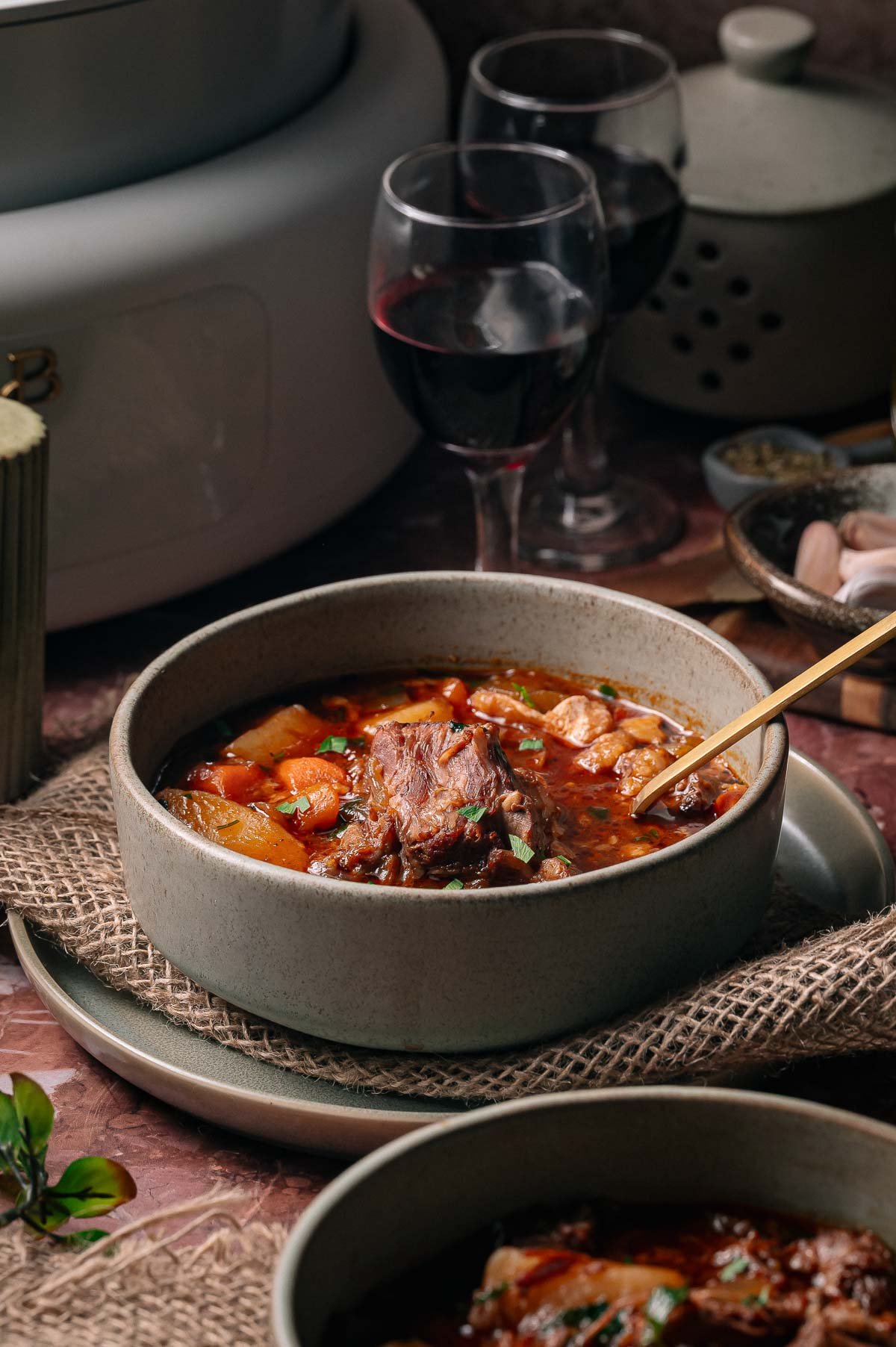 A bowl of oxtail soup with vegetables sits on a plate, accompanied by a spoon, with two glasses of red wine and kitchenware in the background.