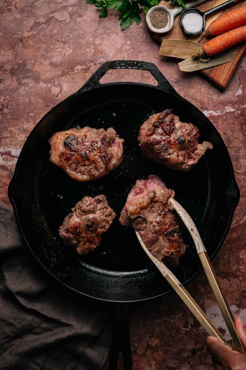 Four browned oxtails cooking in a black cast iron skillet, with metal tongs holding one piece. Vegetables and seasonings are on a wooden board nearby.