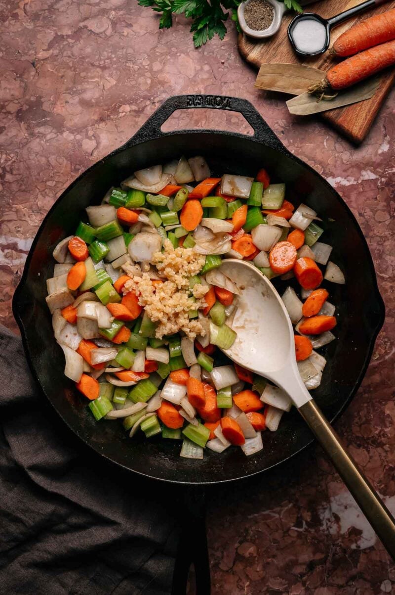 Chopped carrots, celery, onions, and minced garlic are being sautéed in a black skillet with a wooden spoon on a brown marble countertop.