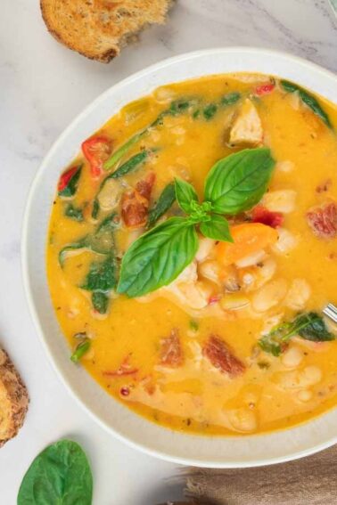 A bowl of creamy soup with beans, greens, and vegetables, garnished with basil, beside toasted bread slices on a marble surface.
