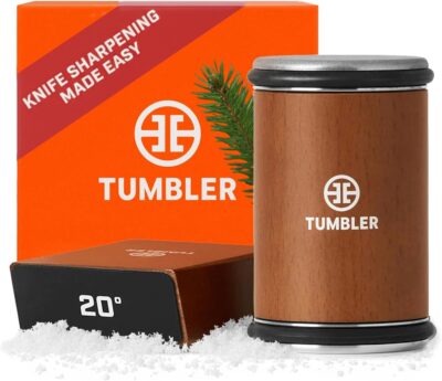 Brown cylindrical knife sharpener labeled "TUMBLER" is shown upright on fake snow beside its orange packaging featuring text "Knife Sharpening Made Easy" and "20ยฐ.