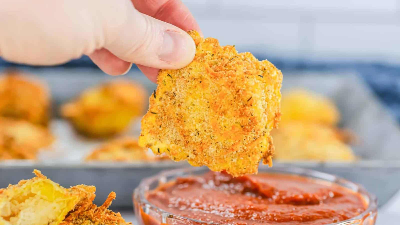 A hand dips a crispy, breaded zucchini chip into a bowl of marinara sauce.