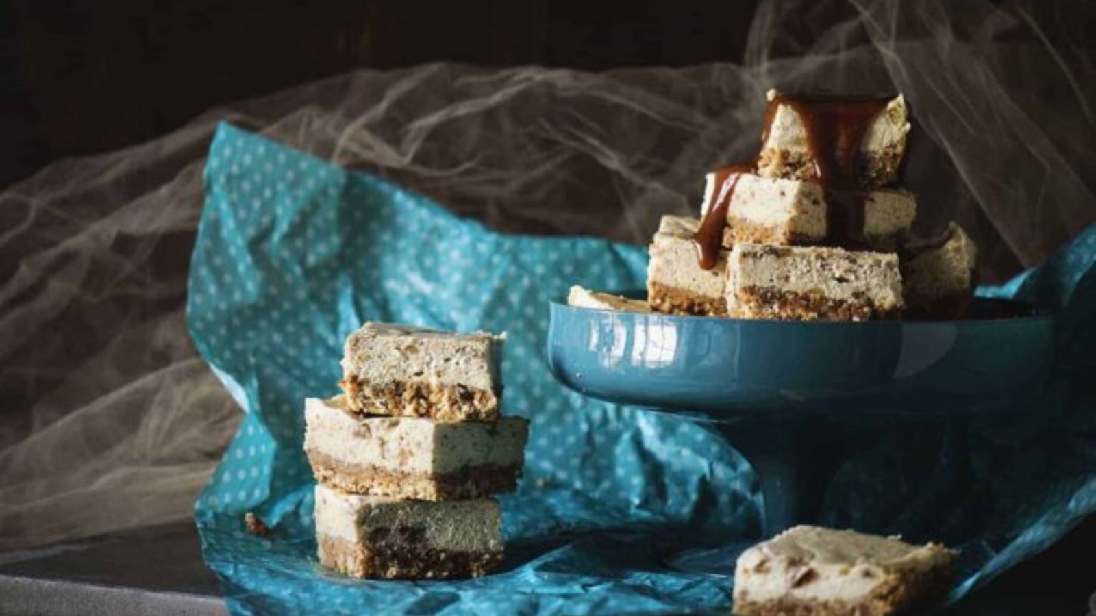 Several cheesecake bars with a crumbly base are stacked on a blue cake stand and blue polka dot fabric, some topped with sauce.