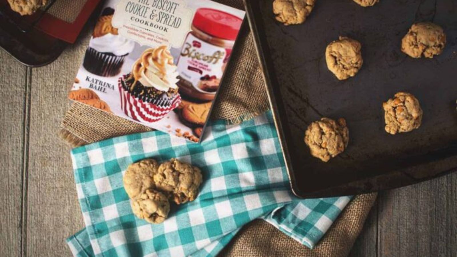 Chocolate chip cookies on a baking tray and a blue plaid cloth, with a cookbook and cookie jars in the background on a wooden surface.