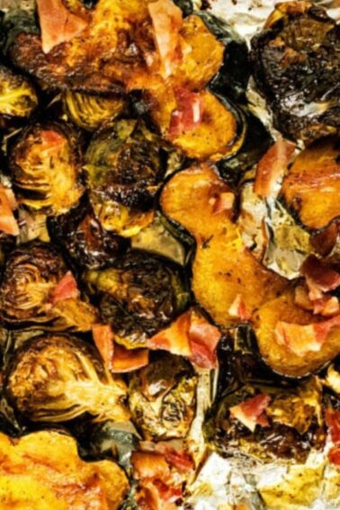 Roasted acorn squash slices and Brussels sprouts with pieces of bacon on a foil-lined baking sheet.