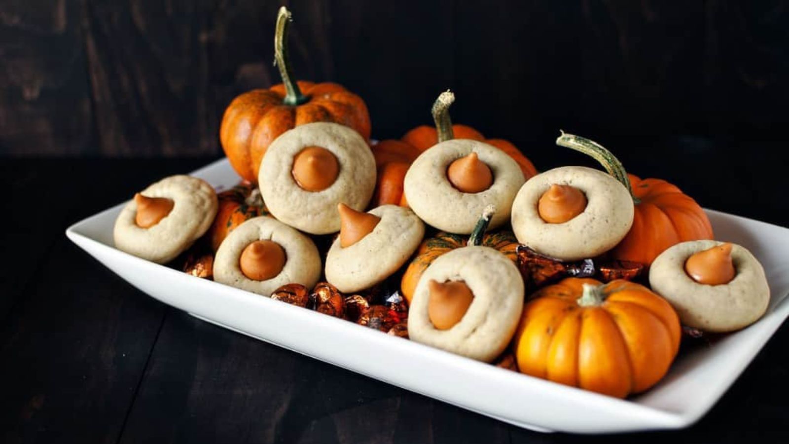A rectangular white plate holds pumpkin-shaped thumbprint cookies with orange candy centers, surrounded by small decorative pumpkins.