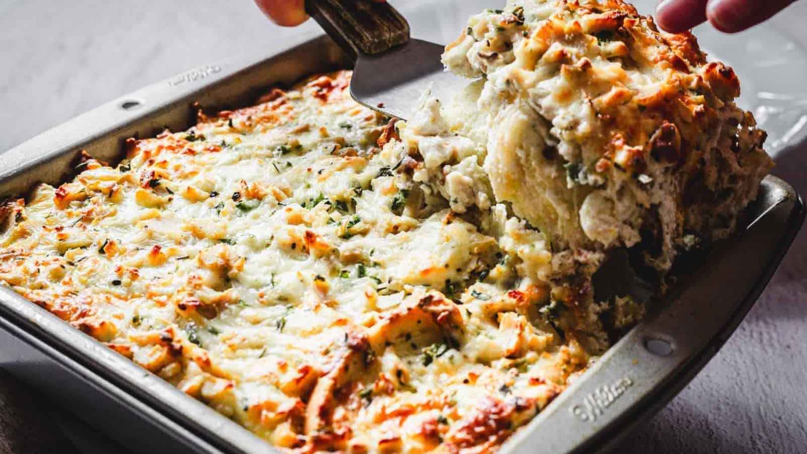 A baked casserole dish filled with a cheesy, golden-brown topping is being scooped with a spatula. The dish appears to contain layers of ingredients beneath the melted cheese, with herbs sprinkled on top for garnish.