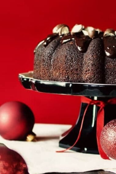 A chocolate bundt cake with chocolate glaze sits on a black cake stand, surrounded by red Christmas ornaments on a white cloth, against a red background.