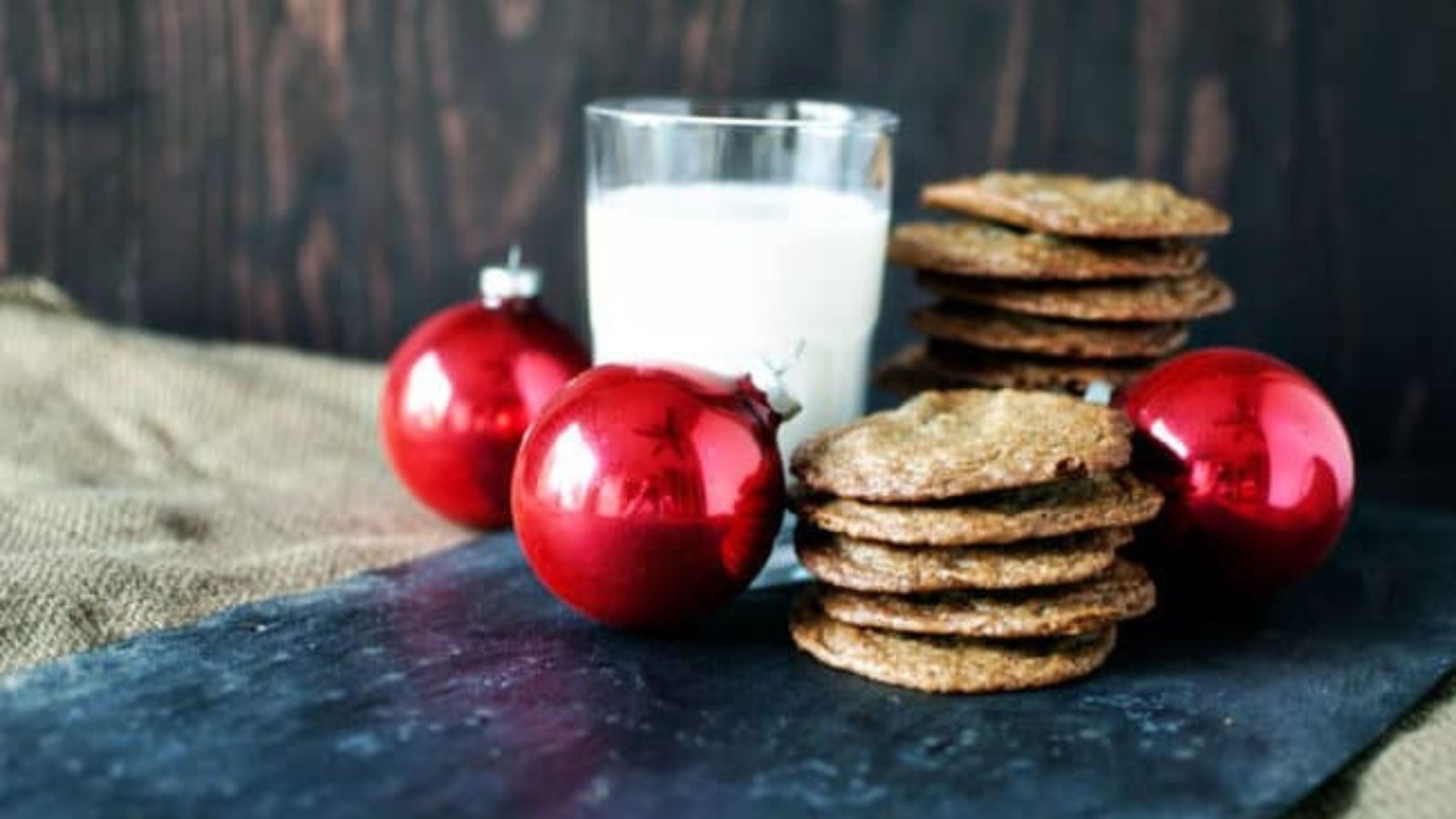 A stack of cookies, two red Christmas ornaments, and a glass of milk set on a dark surface with a burlap cloth and wooden background.