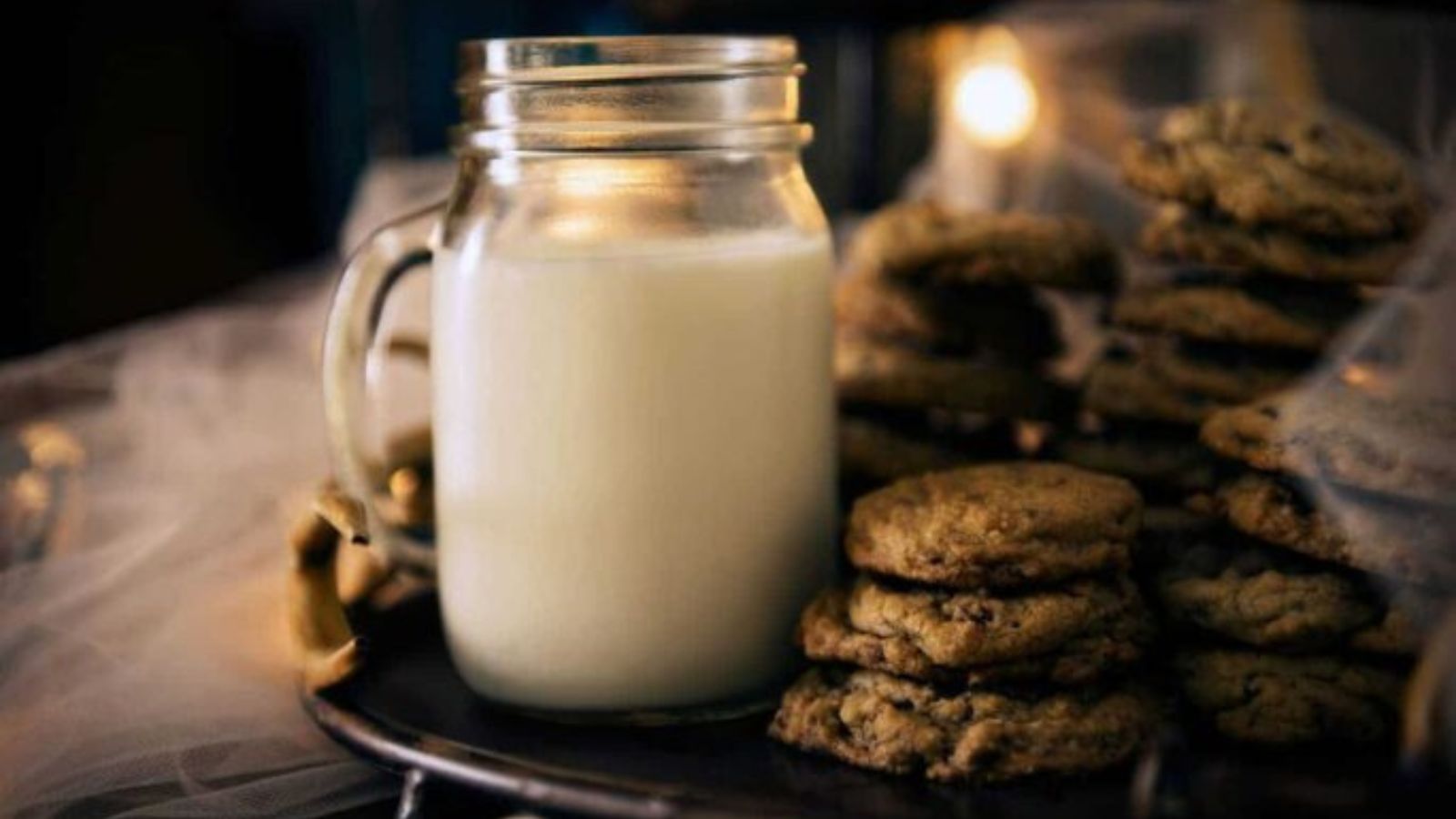 A glass jar filled with milk sits on a tray next to several stacks of chocolate chip cookies, with soft lighting in the background.