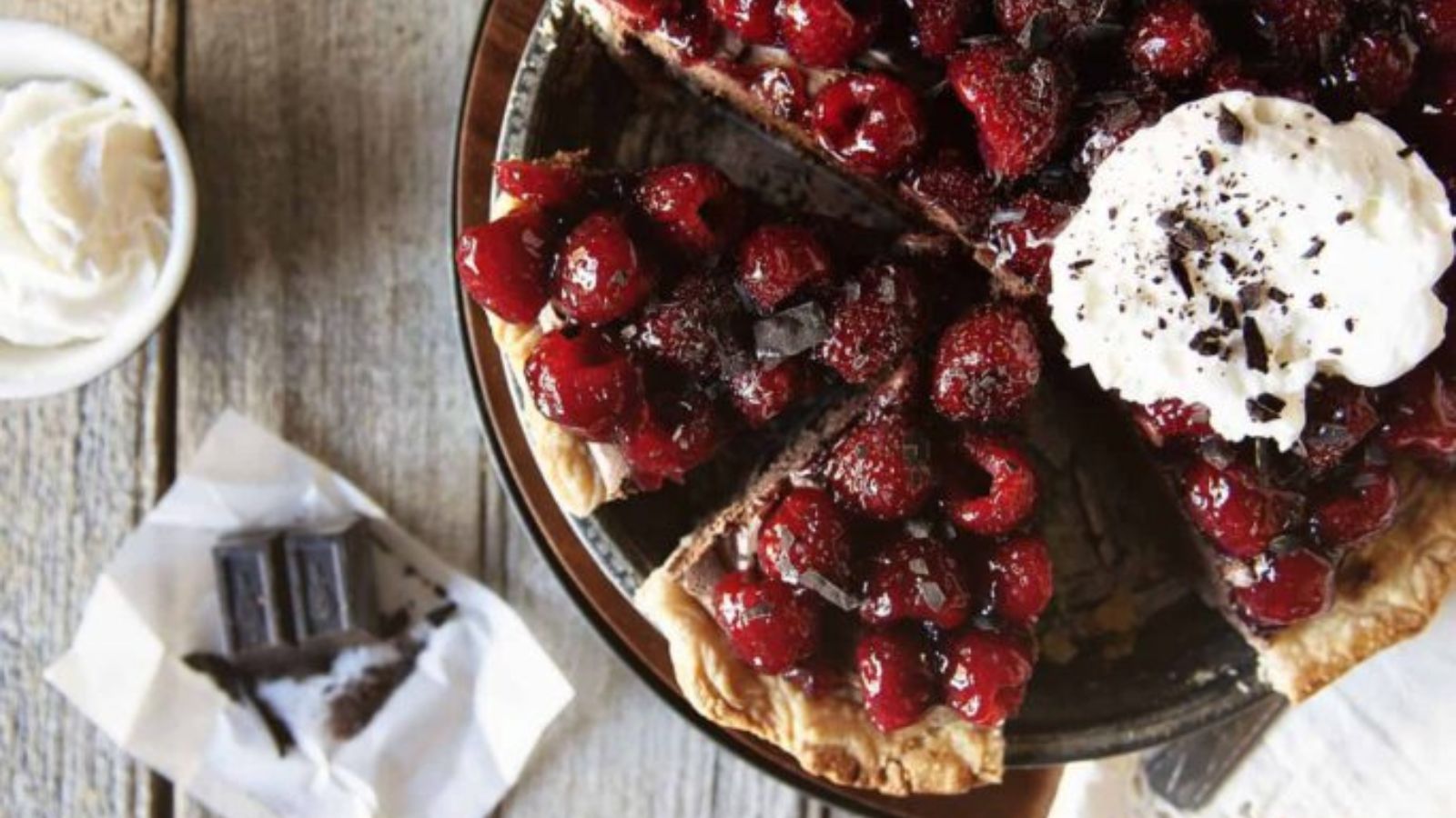 A cherry pie with a whipped cream topping sits on a wooden table, with several slices cut. Nearby are a bar of chocolate and a small bowl of whipped cream.