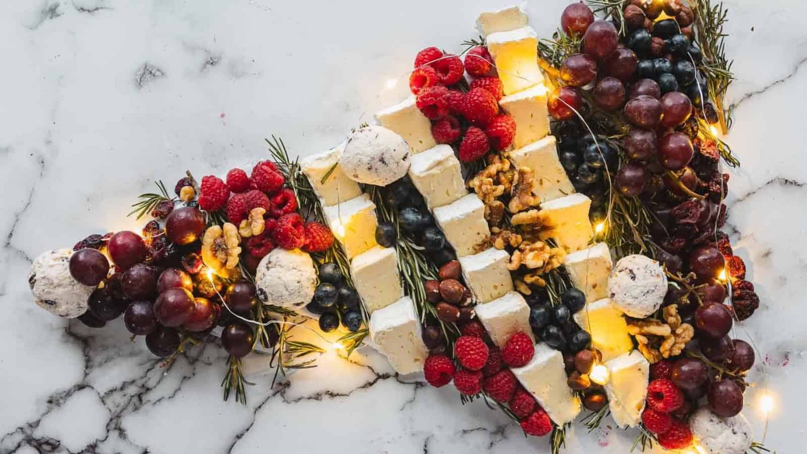 Cheese, grapes, berries, nuts, and herbs arranged in the shape of a Christmas tree, with string lights.
