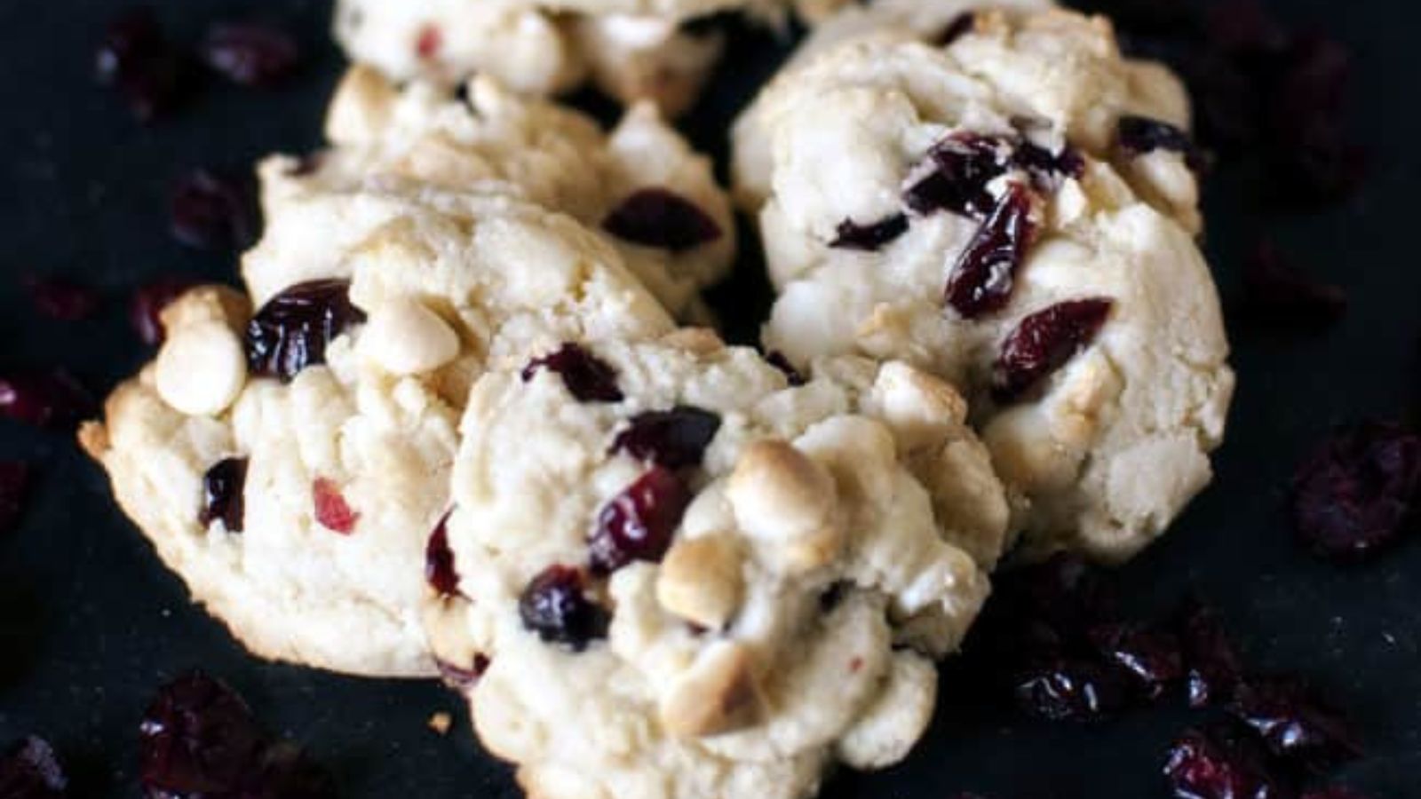 Three white chocolate chip and cranberry cookies on a dark surface with scattered dried cranberries around them.