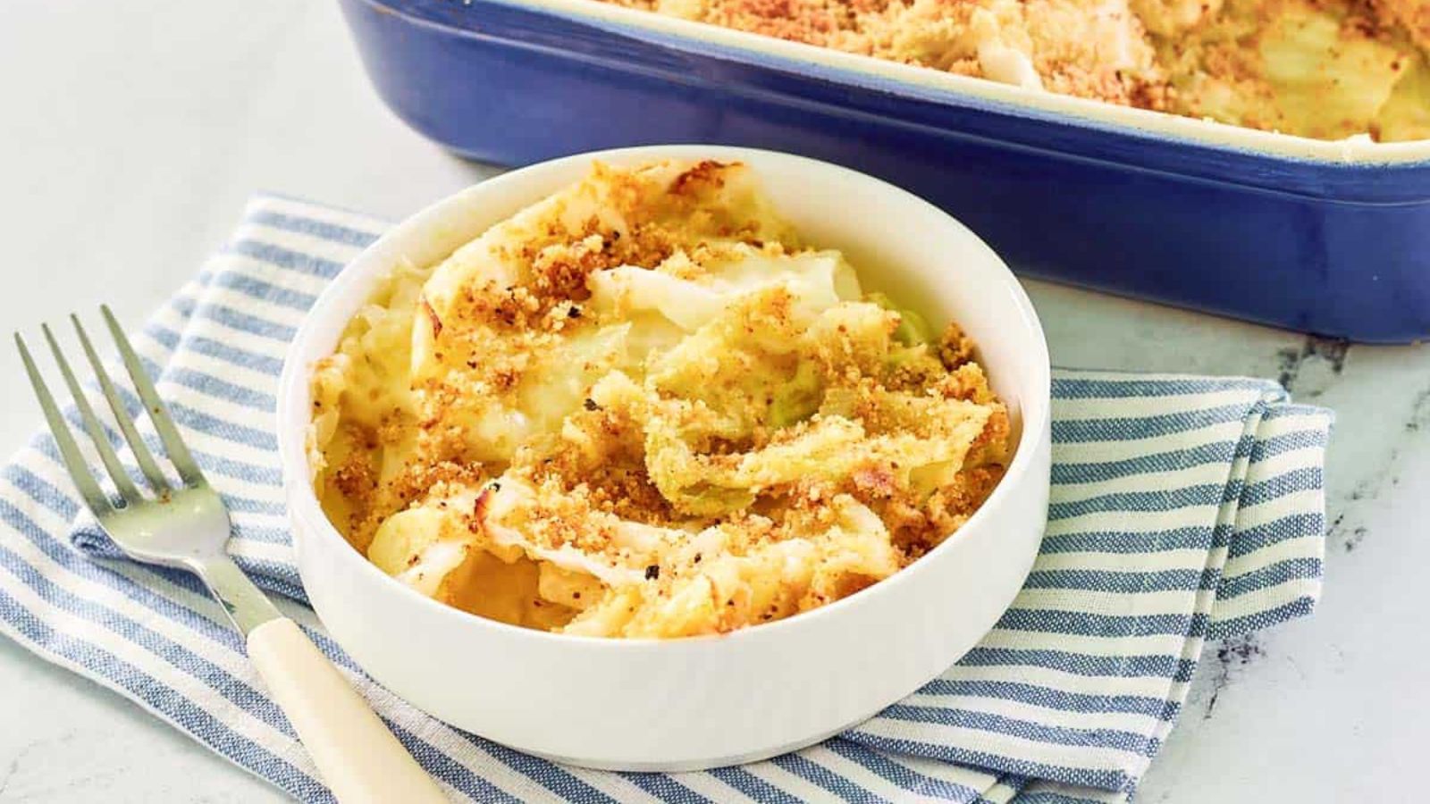 A white bowl of baked mac and cheese with a breadcrumb topping sits on a striped cloth next to a fork; a casserole dish is in the background.
