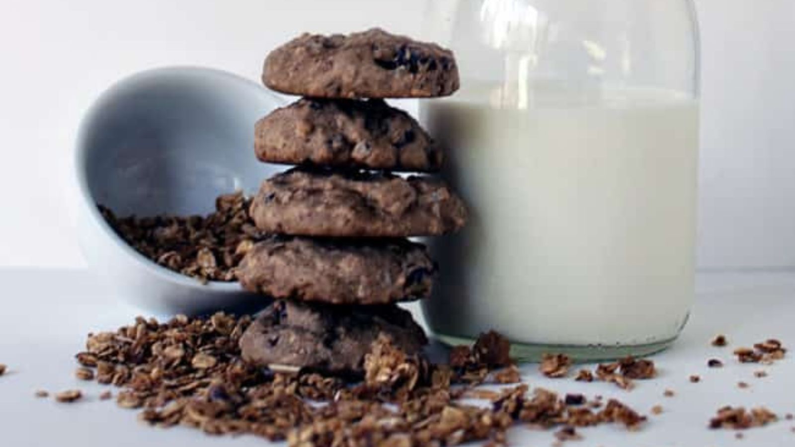 Four chocolate chip cookies stacked on granola, next to a tipped bowl and a glass jar of milk.