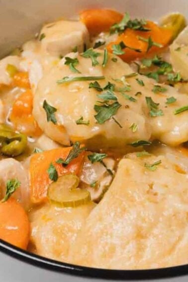 A bowl of chicken and dumplings with carrots, celery, and broth, garnished with chopped parsley. A spoon rests inside the bowl.