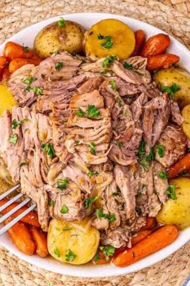 A bowl filled with shredded pot roast, baby potatoes, and carrots, garnished with chopped parsley, sits on a woven placemat with a fork resting on the edge.